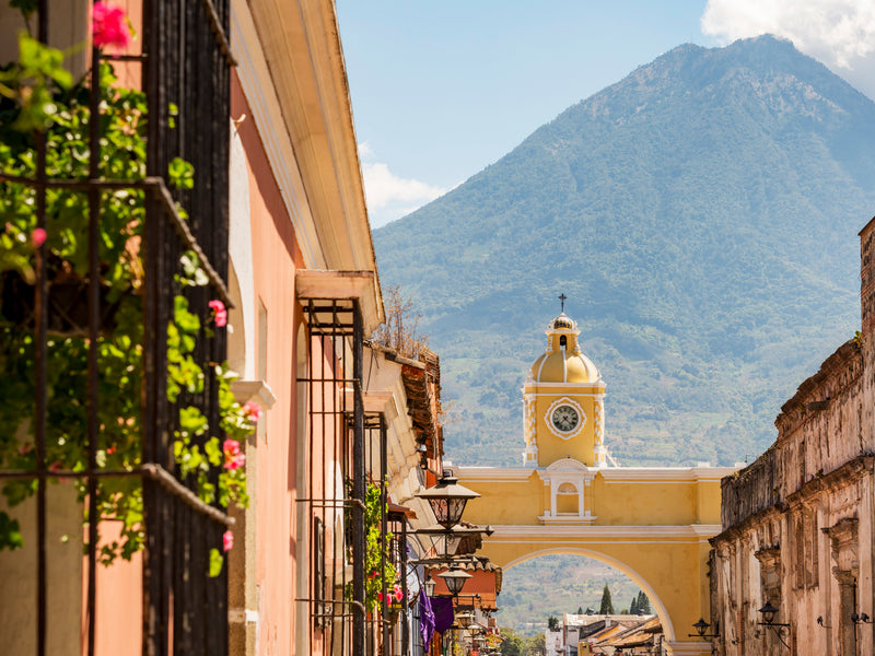 Street scene with a yellow arch and clock tower in Antigua, Guatemala, featuring a mountain in the background; Arco de Santa Catalina
