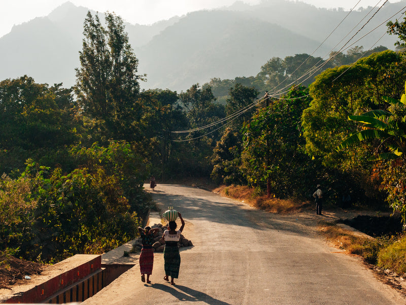 Two people walking on a road surrounded by trees and mountains; Antigua, Guatemala