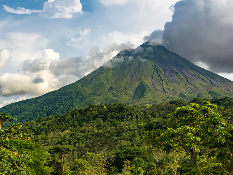Volcano with smoke emerging from the top, surrounded by green trees and a partly cloudy sky; La Fortuna, Arenal, Costa Rica