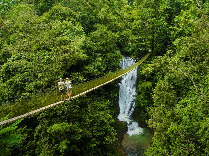 Two people standing on a suspension bridge over a waterfall in a lush green forest; Arenas del Mar Rainmaker suspension bridges Manuel Antonio, Costa Rica