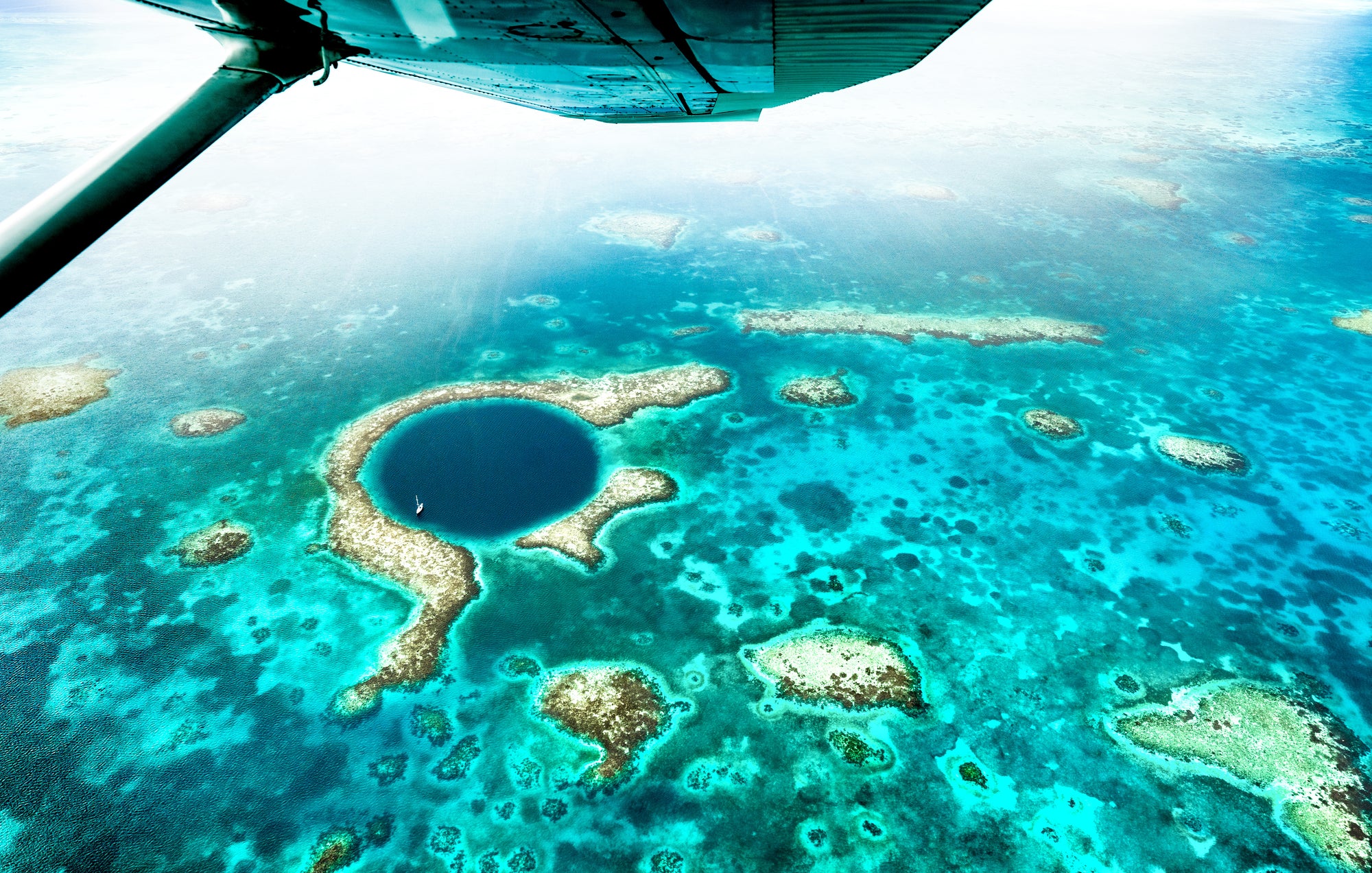 Aerial view of a coral reef with a clear blue water body, likely a sinkhole, in the middle; Belize Blue Hole flyover