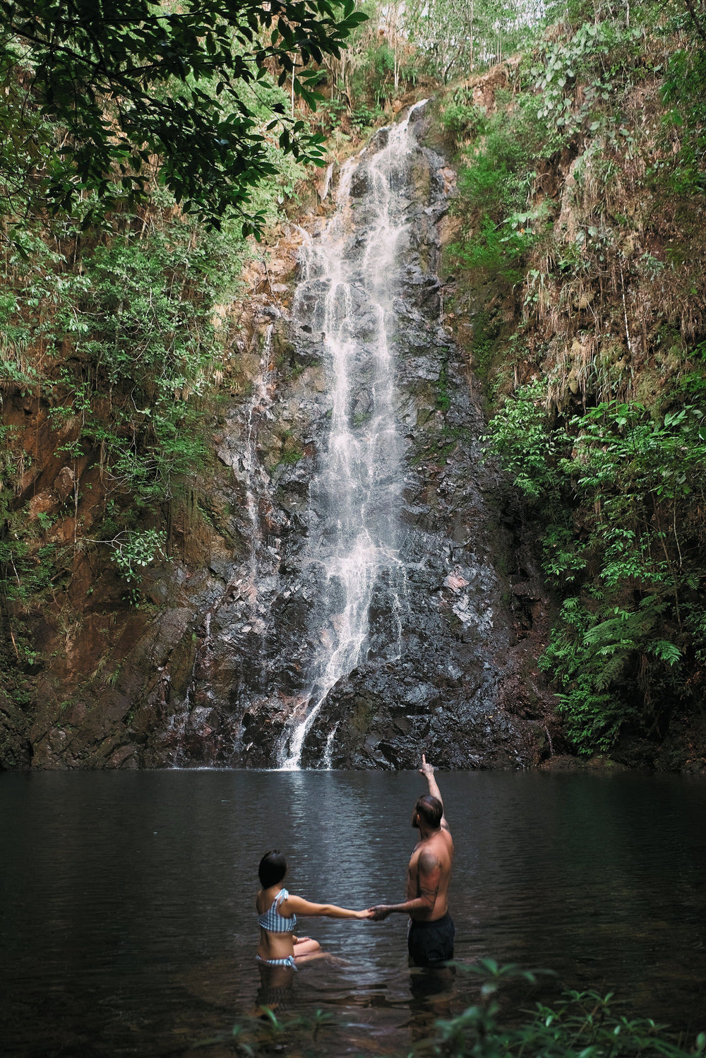 Couple holding hands in front of a waterfall in a lush forest; Hidden Valley Lodge, Mountain Pine Ridge, Belize portrait