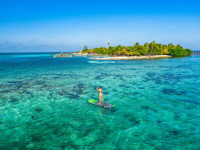 Person paddleboarding in clear blue water with a tropical island in the background, Ray Caye, Belize