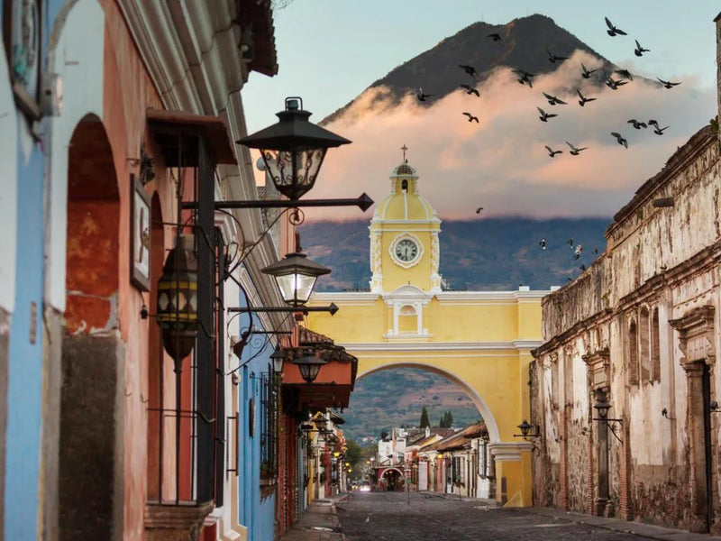 Street scene with a yellow arch and clock tower in the background, mountains in the distance; Antigua, Guatemala