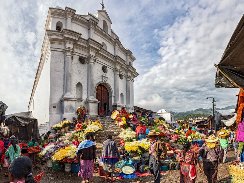 Market scene with people and stalls in front of a large white church under a cloudy sky. Chichicastenango market, Guatemala.