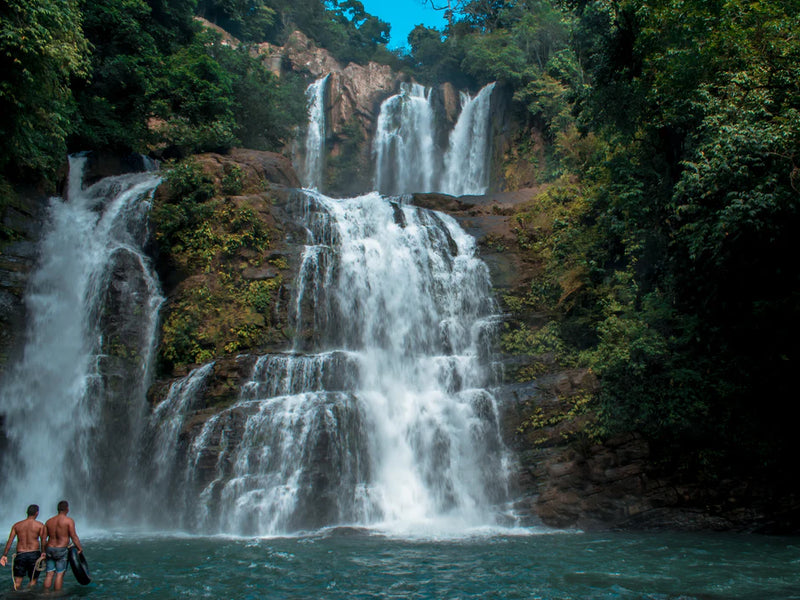 Two people standing at the base of a large waterfall surrounded by lush greenery; Nauyaca Falls, Dominical, Costa Rica