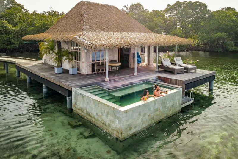 Floating water villa with a thatched roof on a body of water, surrounded by greenery; Bocas del Toro Nayara Panama