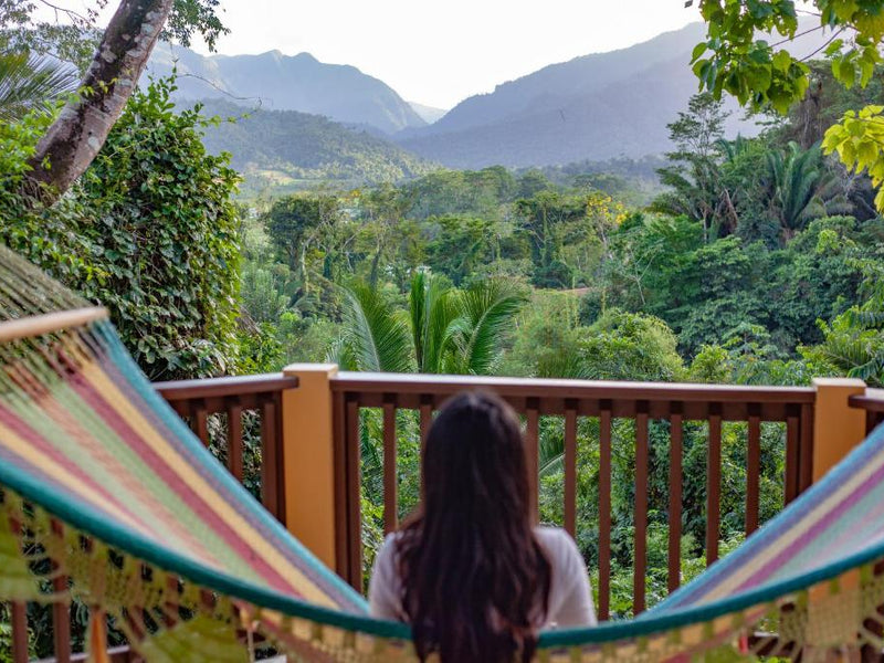 Person sitting in a colorful hammock overlooking a lush green landscape with mountains. Sleeping Giant Lodge, Belmopan. Belize.
