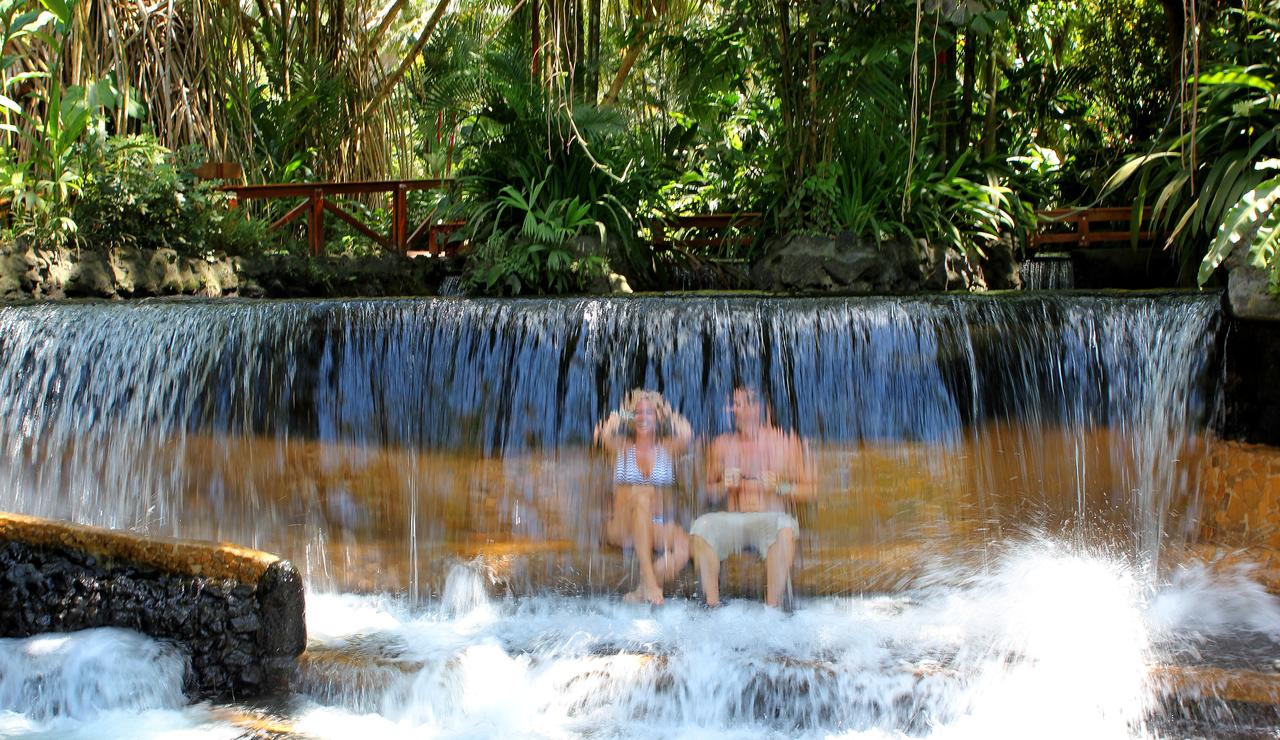 Two people sitting by a waterfall in a lush, green forest setting; Tabacon hot springs, Arenal, La Fortuna, Costa Rica